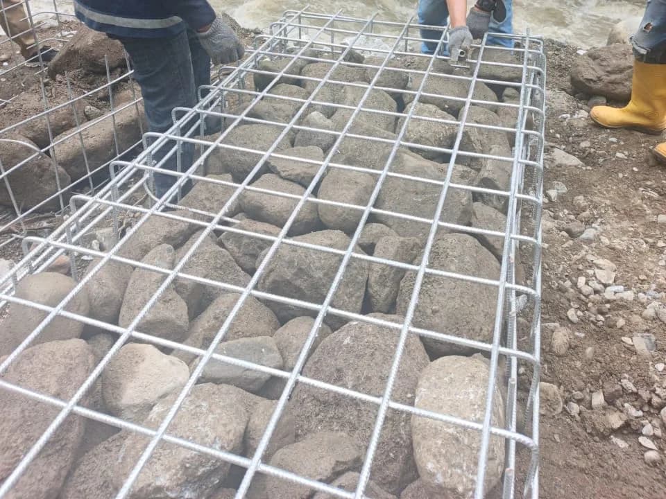 Panel bronjong kawat terpasang berisi batu kali di tepi sungai, dengan pekerja bersarung tangan merapikan sambungan panel.